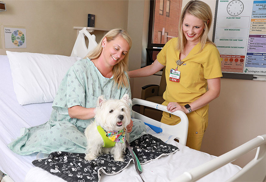 A smiling nurse and patient petting a dog.